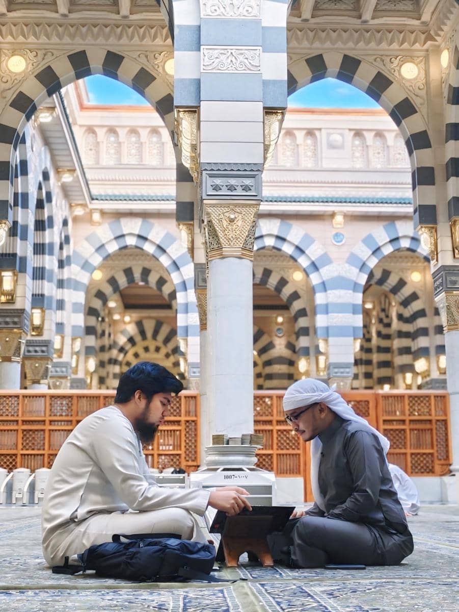 Dua pria mengenakan pakaian muslim duduk bersila di dalam masjid megah dengan arsitektur berpilar dan kubah berwarna biru keabu-abuan.