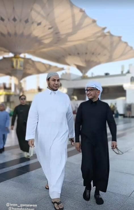 Dua pria mengenakan jubah putih dan hitam berjalan bersama di area Masjid Nabawi, dengan latar payung besar khas Madinah.