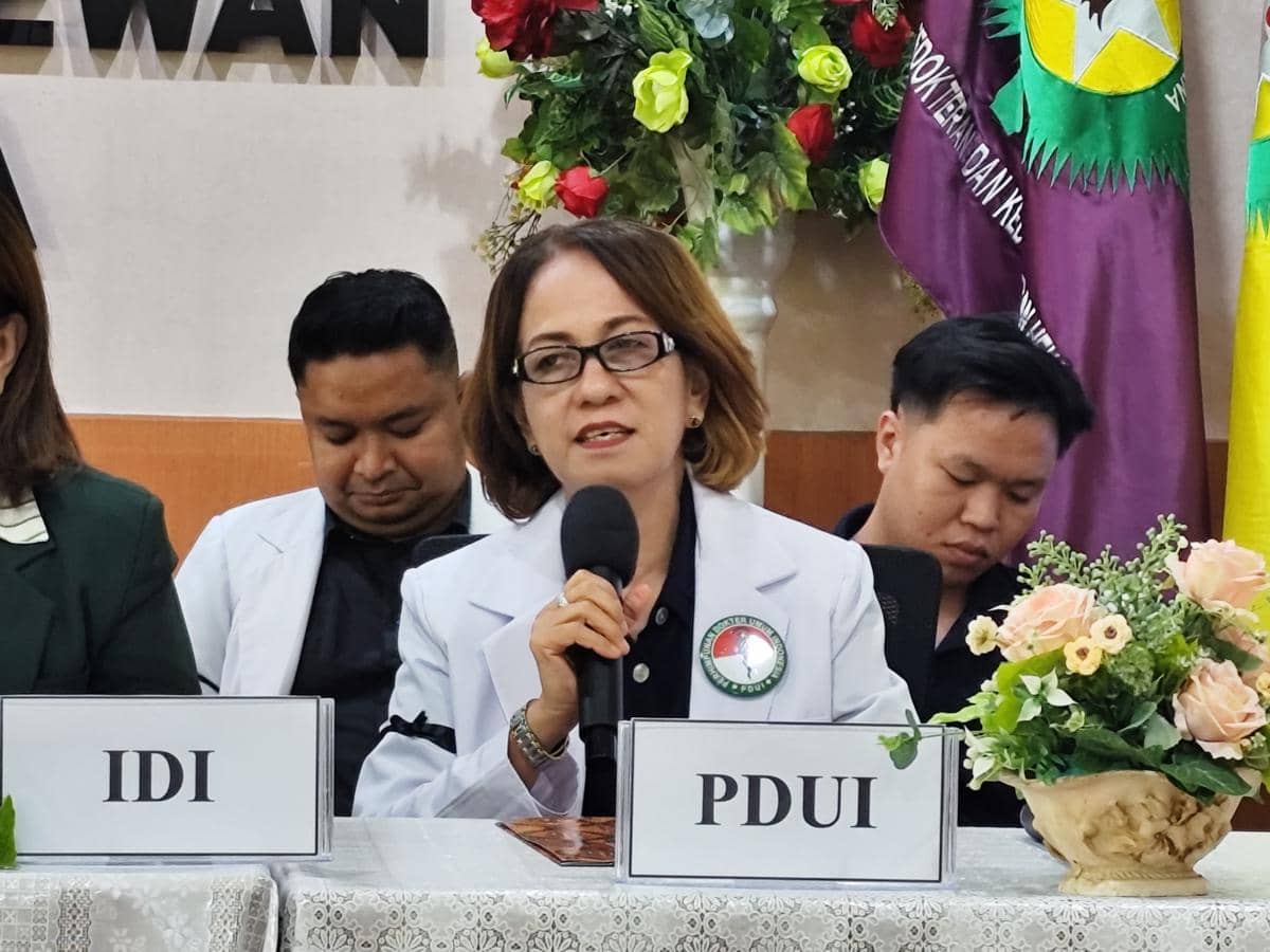 Seorang perempuan mengenakan jas dokter berbicara memegang mikrofon di meja konferensi dengan papan nama PDUI di depannya.
