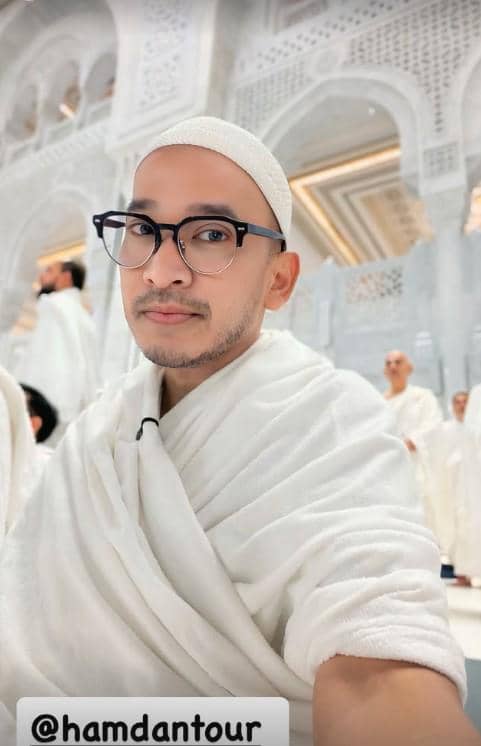 Seorang pria mengenakan pakaian ihram berwarna putih dan peci putih sedang berada di area masjid dengan arsitektur megah berwarna putih.