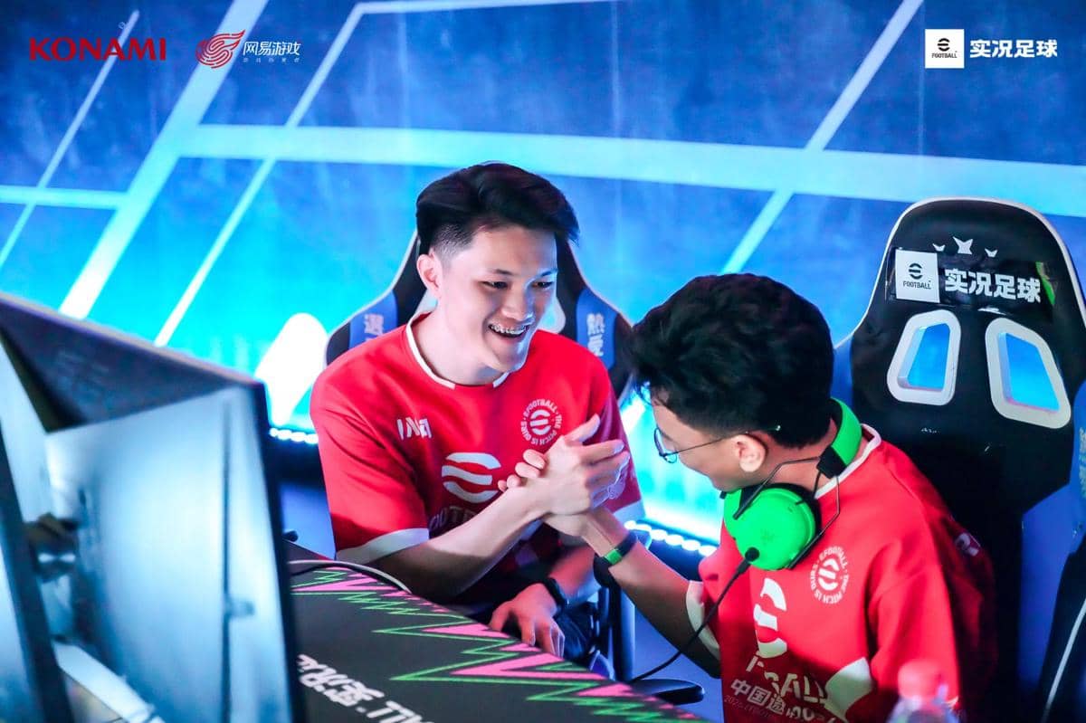 Dua atlet eFootball Indonesia mengenakan jersey merah saling berjabatan tangan dengan ekspresi gembira di depan komputer saat turnamen.
