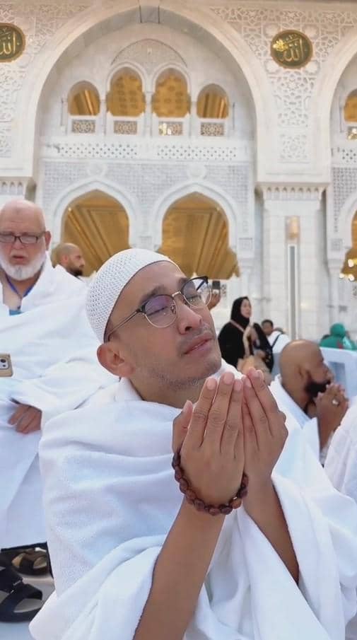 Seorang pria mengenakan pakaian ihram putih dan peci sedang berdoa dengan khusyuk di area masjid dengan arsitektur megah berwarna putih.