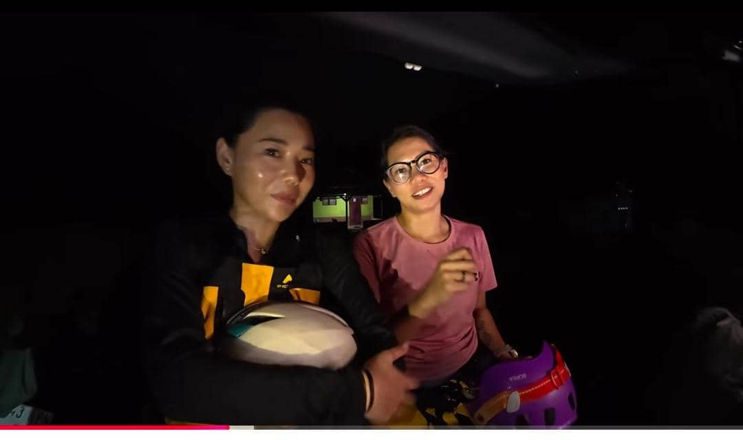 Dua perempuan berada di dalam gua dengan pencahayaan minim, salah satunya memegang helm dan mengenakan pakaian olahraga.