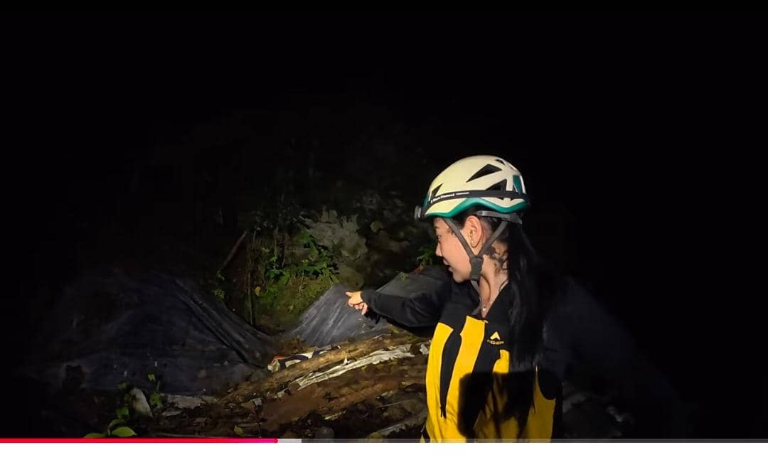 Seorang perempuan mengenakan helm dan jaket kuning hitam menunjuk ke arah pintu masuk Gua Bojong Nangka di malam hari.