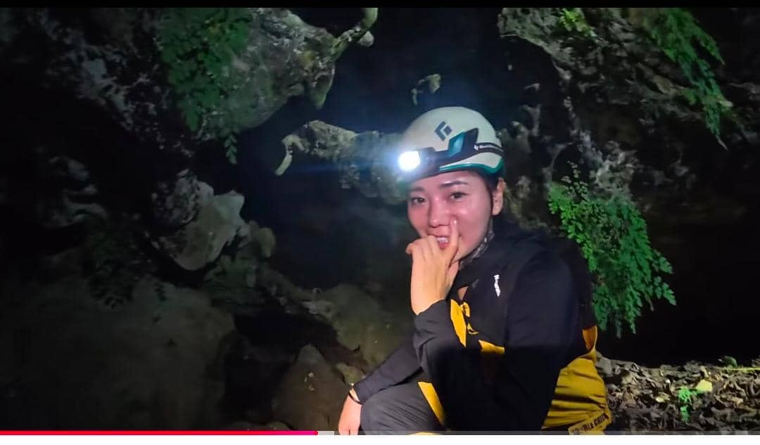 Seorang perempuan mengenakan helm dengan lampu kepala dan pakaian pendakian sedang berada di dalam Gua Bojong Nangka yang gelap.