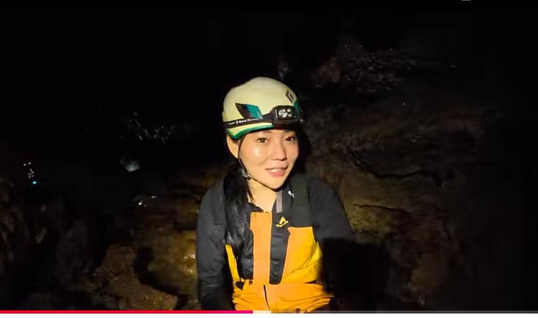 Seorang perempuan mengenakan helm dan pakaian pelindung kuning hitam sedang berada di dalam Gua Bojong Nangka yang gelap.