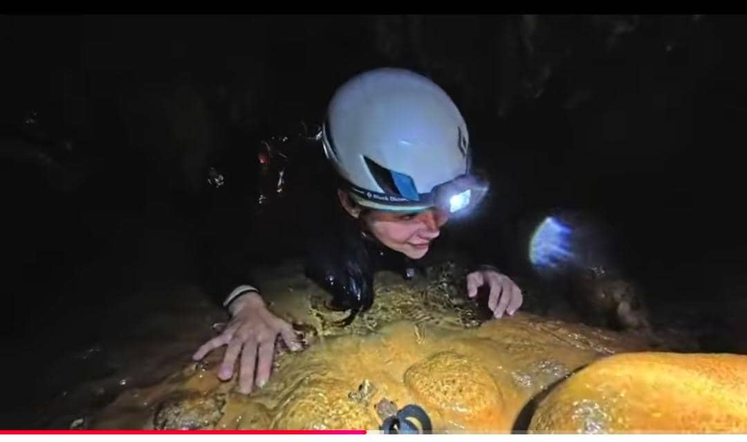 Seorang perempuan mengenakan helm keselamatan dan lampu kepala sedang merangkak di dalam Gua Bojong Nangka yang gelap dan lembap.