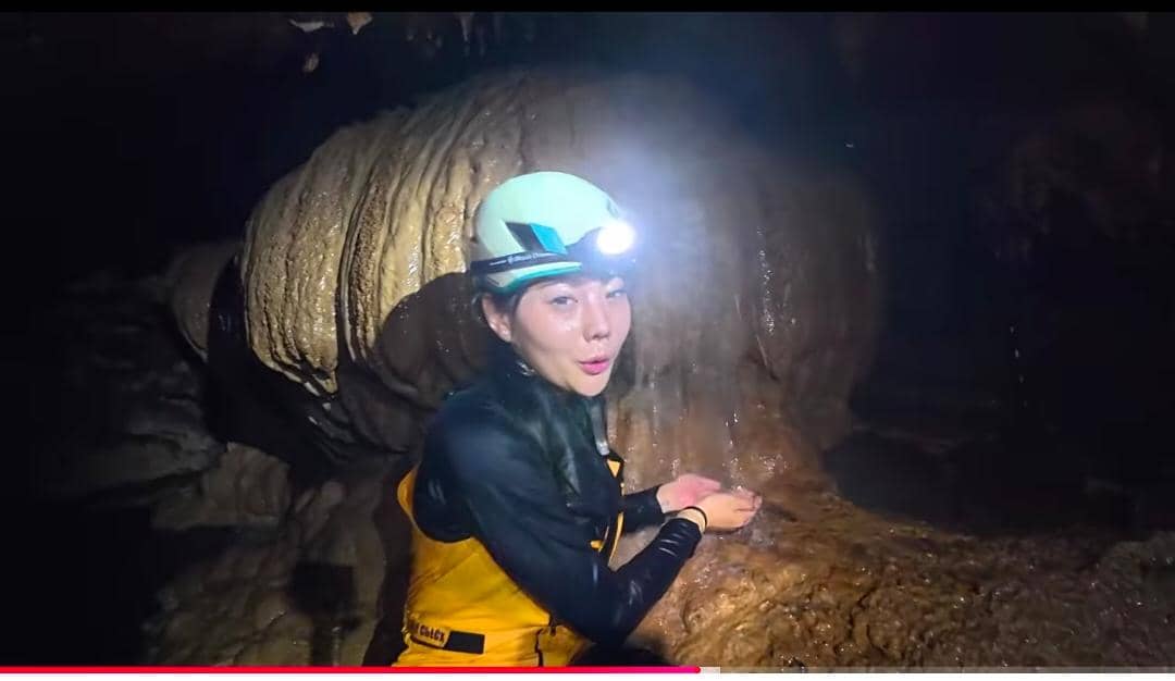 Seorang perempuan mengenakan helm dengan lampu kepala dan pakaian pelindung kuning sedang berada di dalam Gua Bojong Nangka yang lembap.