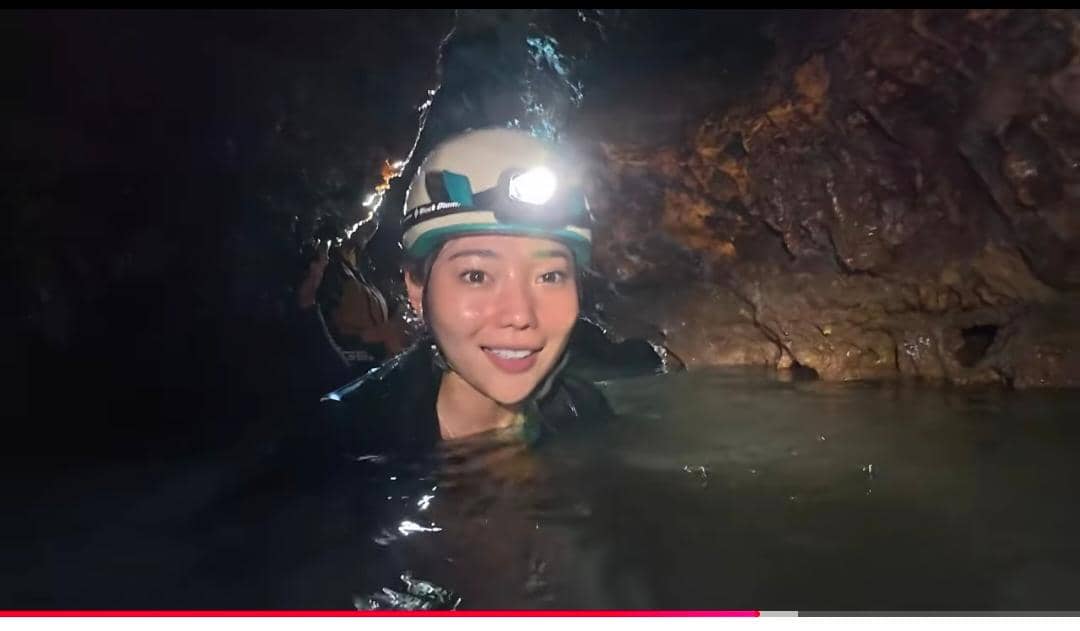 Seorang perempuan mengenakan helm dengan lampu kepala berada di dalam gua berair, tampak tersenyum di tengah pencahayaan redup.