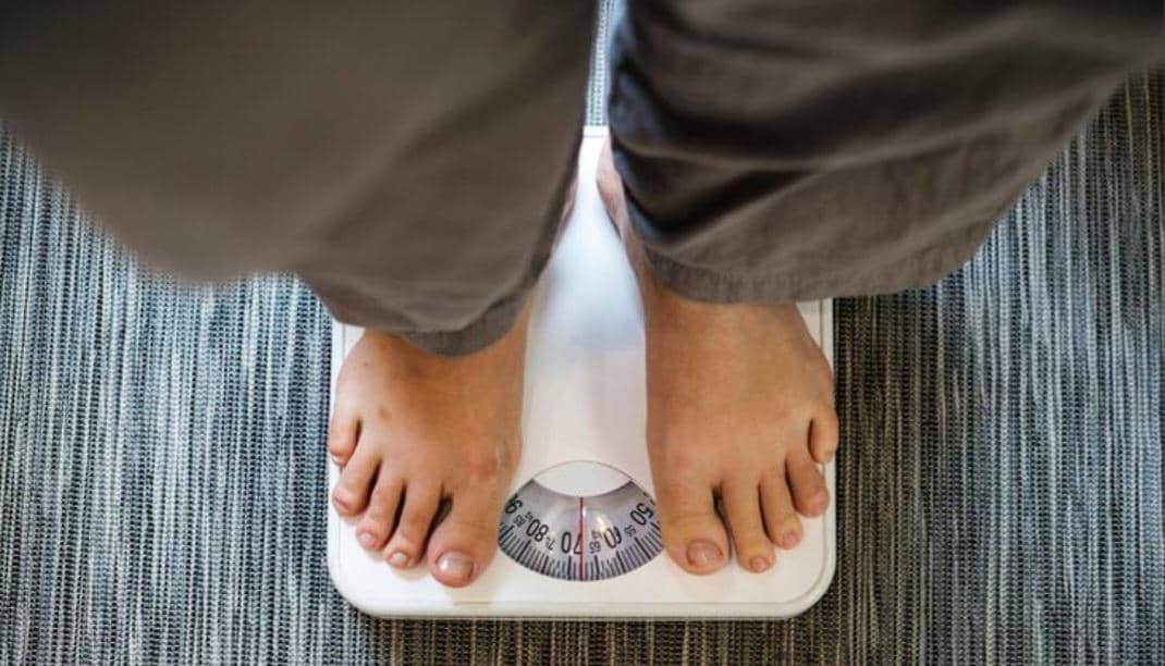 Seseorang berdiri di atas timbangan analog dengan jarum penunjuk berat badan di atas permukaan karpet abu-abu.