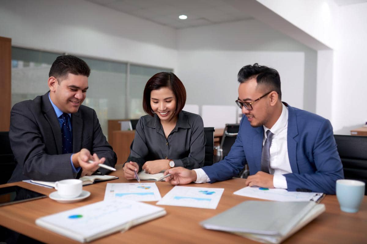 Tiga profesional berdiskusi di ruang rapat sambil meninjau dokumen dan grafik bisnis di atas meja kantor modern.