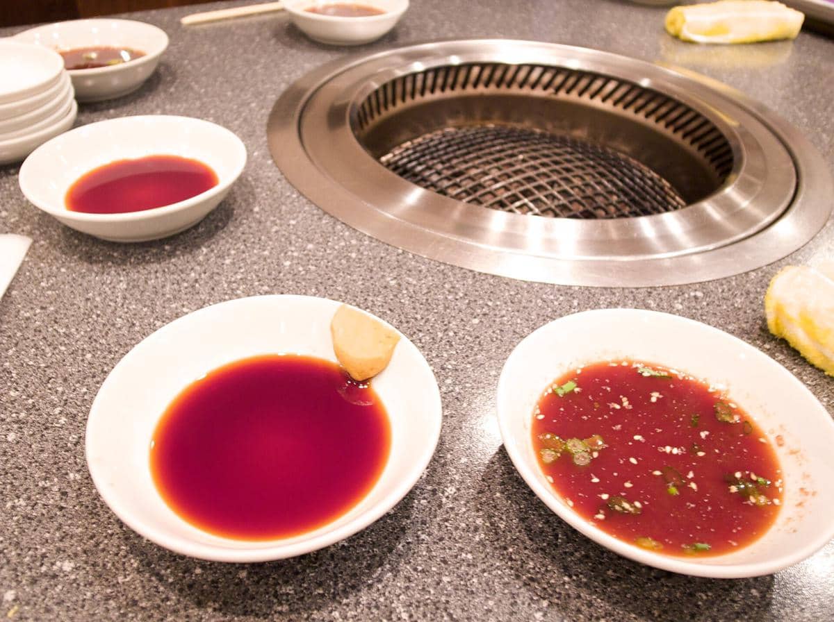Beberapa mangkuk saus yakiniku tare berwarna cokelat kemerahan di atas meja dekat panggangan logam di restoran Jepang.