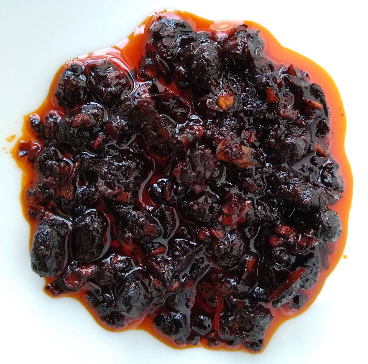Chilli oil berwarna merah gelap dengan potongan cabai dan minyak mengilap tersaji di atas piring putih.