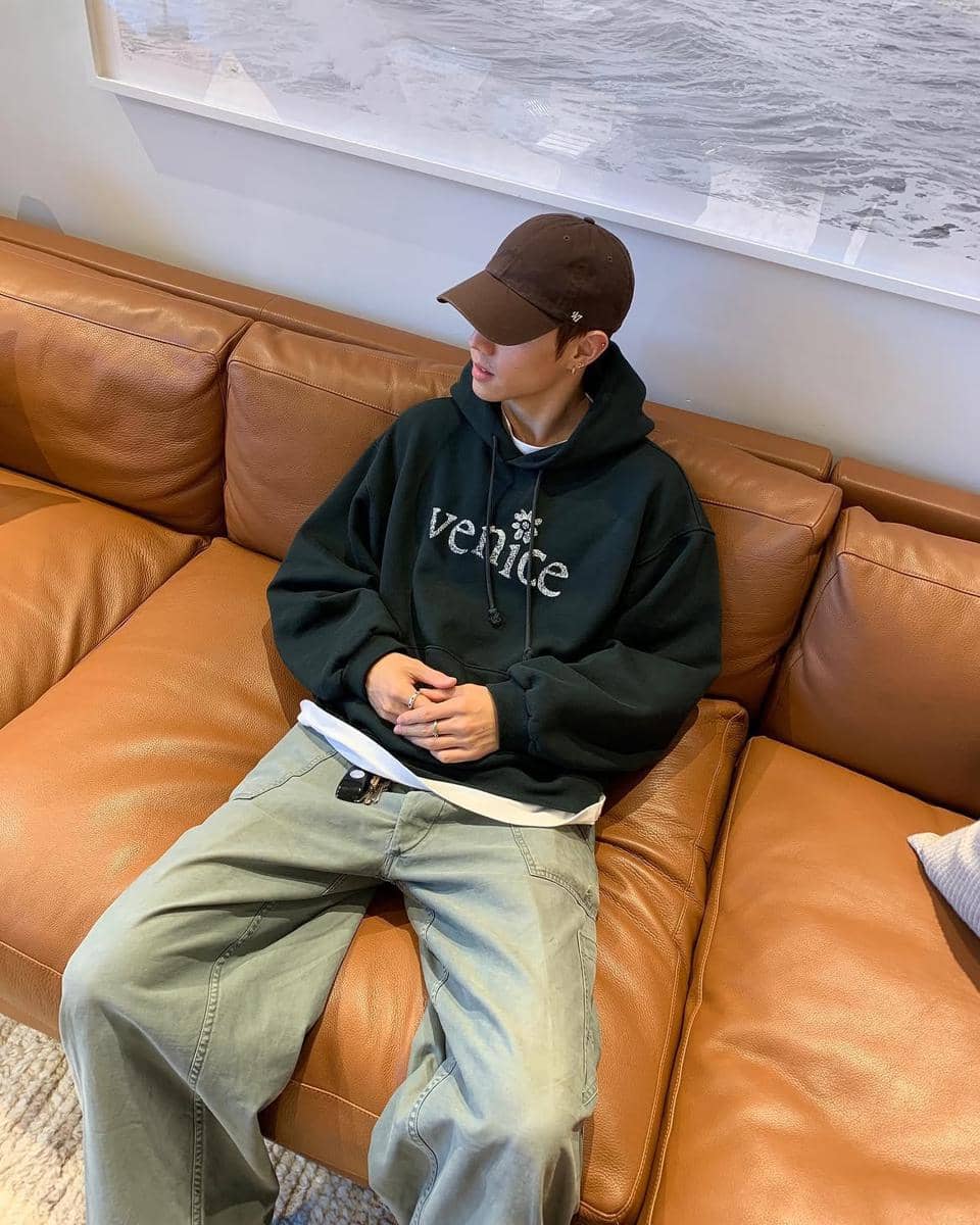 Seorang pria mengenakan hoodie hijau tua bertuliskan 'Venice', celana longgar abu-abu, dan topi cokelat duduk santai di sofa kulit cokelat.