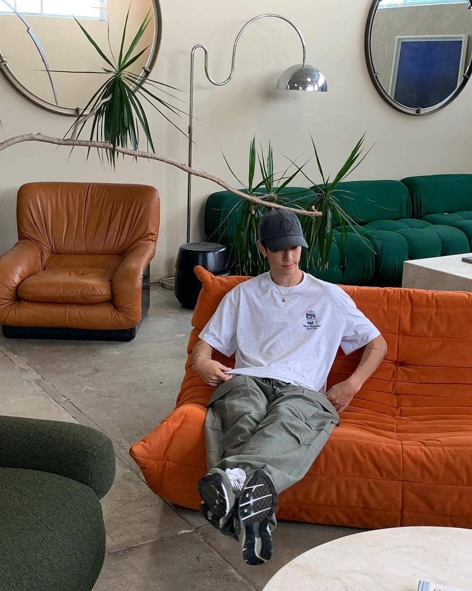 Seorang pria mengenakan kaus oversize abu-abu, celana kargo hijau, dan topi hitam duduk santai di sofa oranye di ruang bergaya modern.