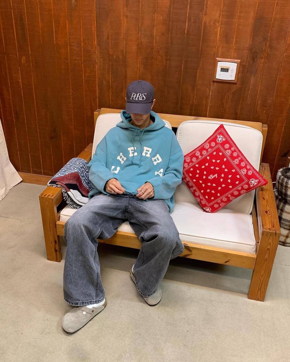 Pria mengenakan hoodie biru oversize bertuliskan 'CHERRY', celana jeans longgar, dan topi hitam duduk di sofa kayu dengan bantal merah.