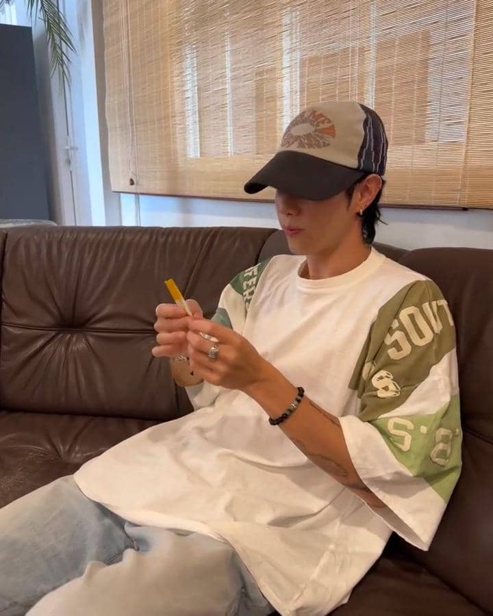 Pria mengenakan kaus oversize putih dengan lengan hijau dan topi baseball duduk di sofa kulit sambil memegang pensil kuning.