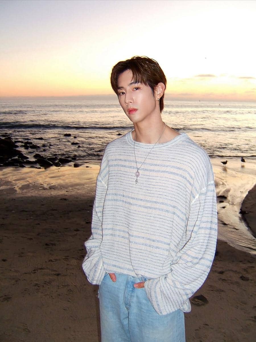Seorang pria mengenakan sweater oversize bergaris dan celana jeans longgar berdiri di tepi pantai saat matahari terbenam.