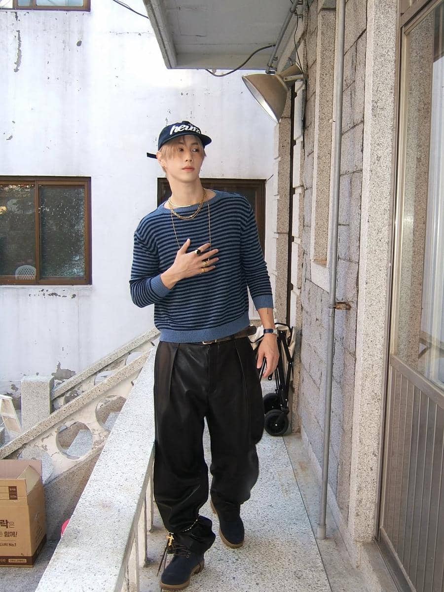 Pria mengenakan sweater garis-garis biru oversize, celana kulit longgar hitam, topi hitam, dan sepatu boots gelap di area balkon bangunan.