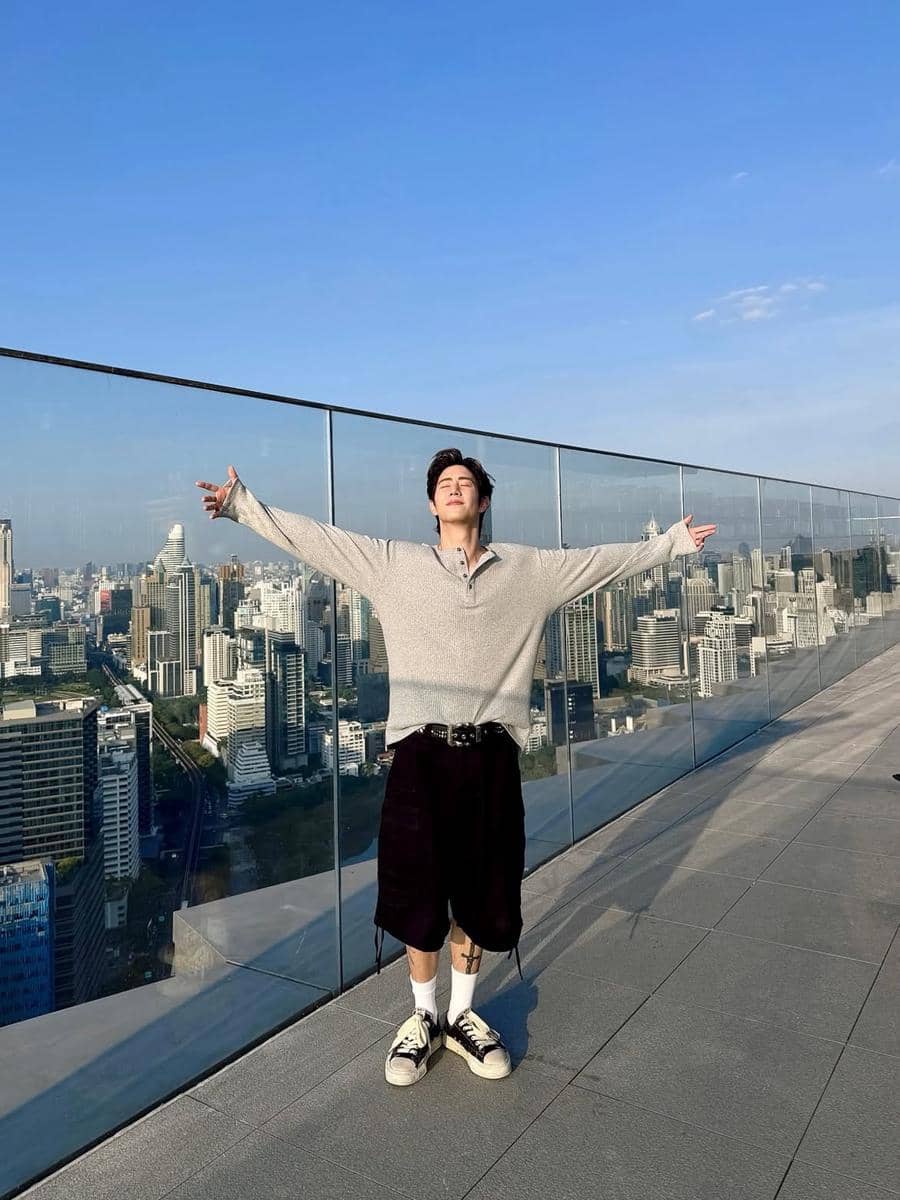 Seorang pria mengenakan sweater oversize abu-abu dan celana pendek hitam berpose dengan tangan terbuka di atap gedung dengan latar kota modern.