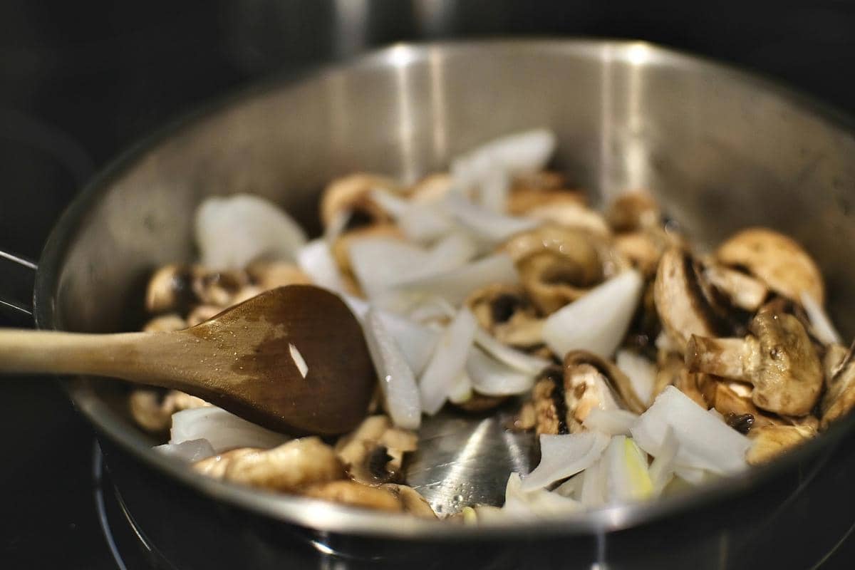 Potongan bawang bombai dan jamur sedang ditumis dalam wajan stainless steel dengan sendok kayu di atas kompor.