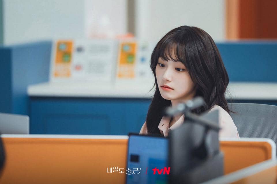 Seorang wanita berambut panjang duduk di meja kantor dengan ekspresi serius, mengenakan kemeja terang di lingkungan kerja modern.