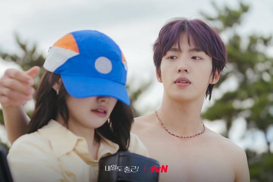 Adegan dua karakter dalam drama Korea See You at Work Tomorrow, seorang pria tanpa baju berbicara dengan wanita ber topi biru di luar ruangan.