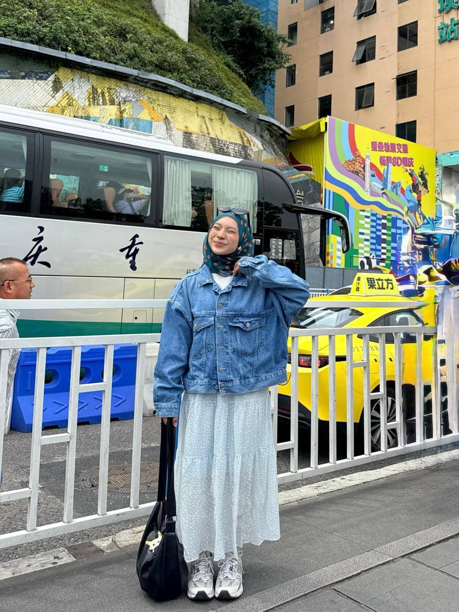 Seorang wanita berhijab mengenakan jaket denim, rok panjang berwarna terang, dan sepatu sneakers berpose di trotoar kota dengan latar bus dan taksi.