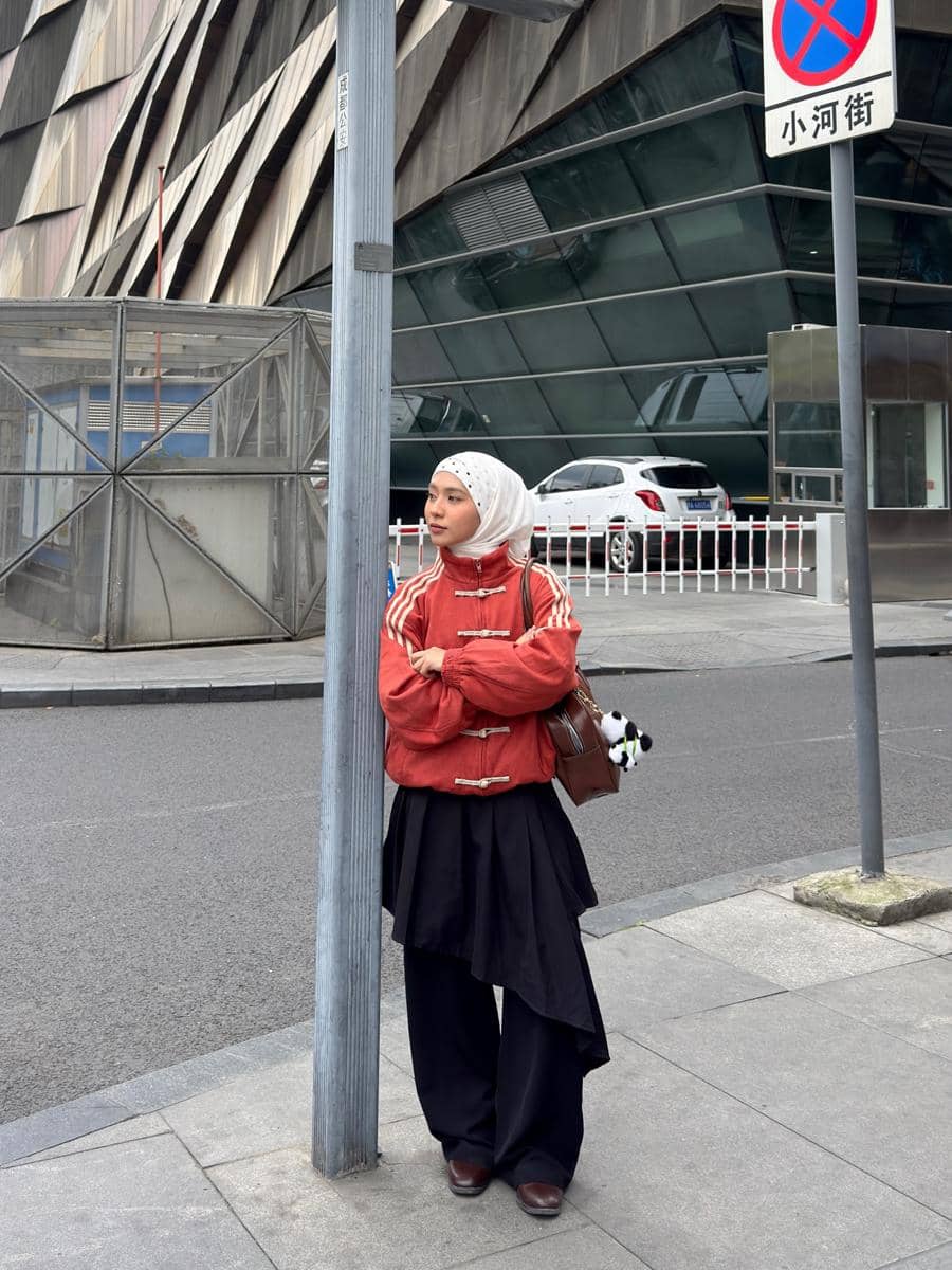 Perempuan berhijab mengenakan jaket merah dan rok hitam berdiri di trotoar kota dengan latar gedung modern dan mobil putih.