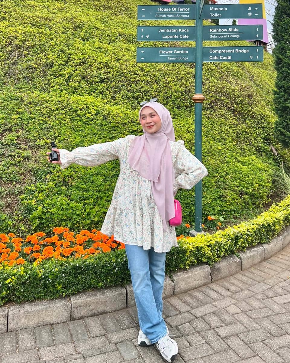 Perempuan berhijab berpose santai di taman dengan latar papan petunjuk arah wisata dan deretan bunga oranye yang rapi.