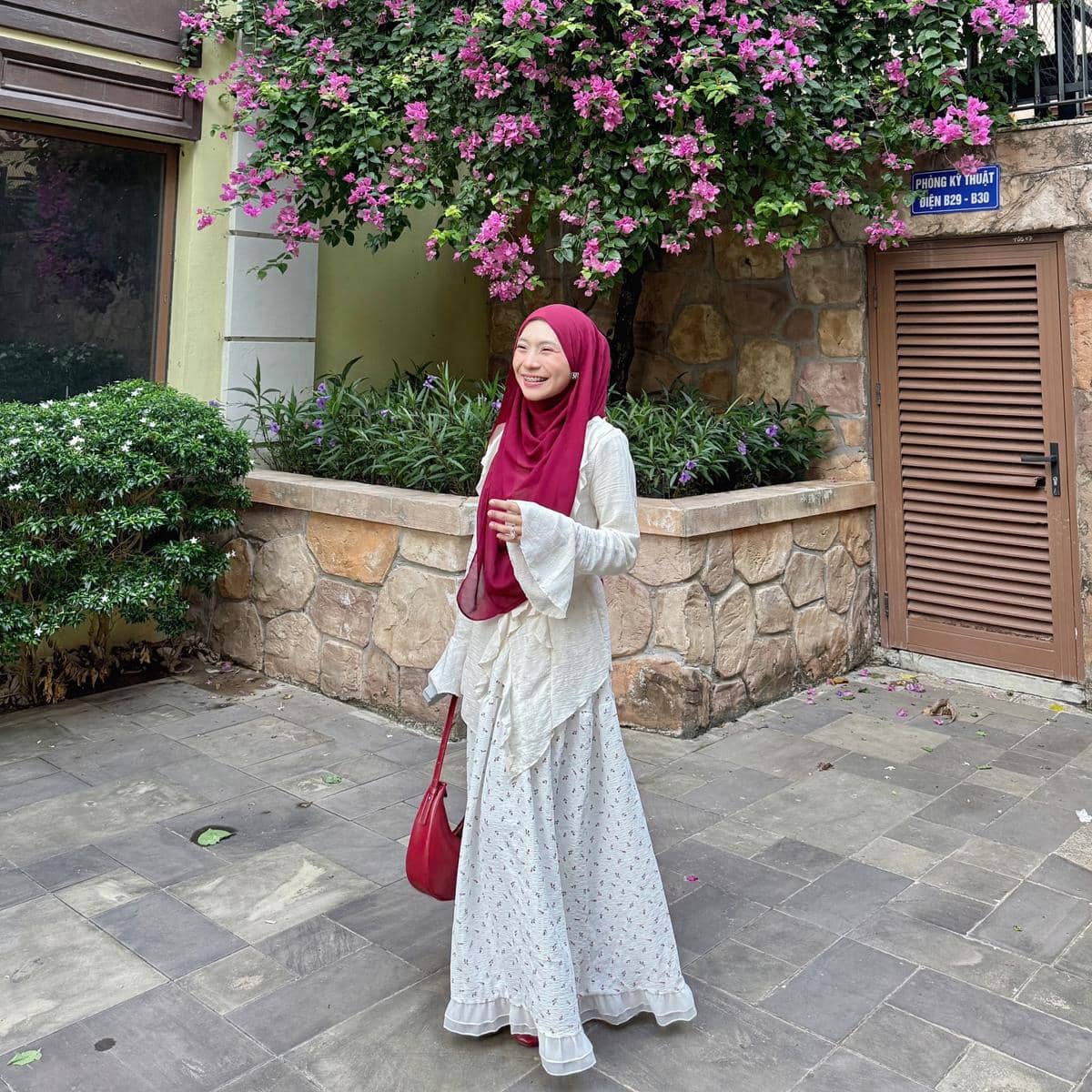 Seorang wanita berhijab merah marun mengenakan dress putih bermotif kecil berjalan di area taman dengan bunga bougenville ungu.