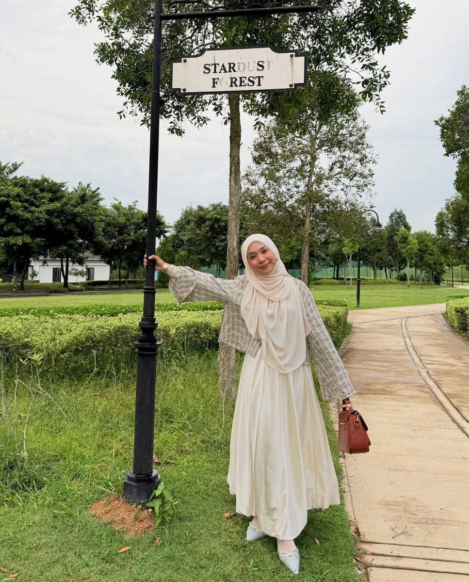 Seorang wanita berhijab berpose santai di taman hijau dengan papan bertuliskan Stardust Forest sambil mengenakan gaun krem dan tas cokelat.
