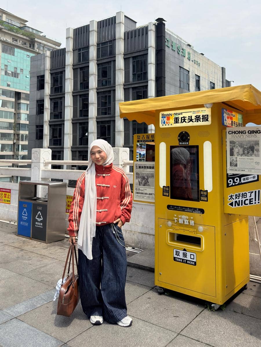 Seorang wanita berhijab mengenakan jaket merah dan celana panjang garis-garis biru berpose di dekat mesin foto kuning di area perkotaan.