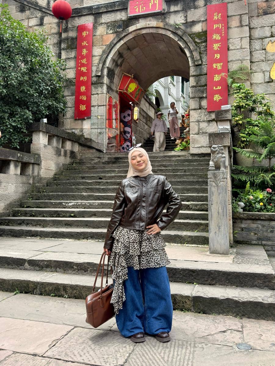 Perempuan berhijab berpose di depan gerbang batu bergaya oriental dengan hiasan lentera merah, mengenakan jaket kulit cokelat dan celana denim lebar.