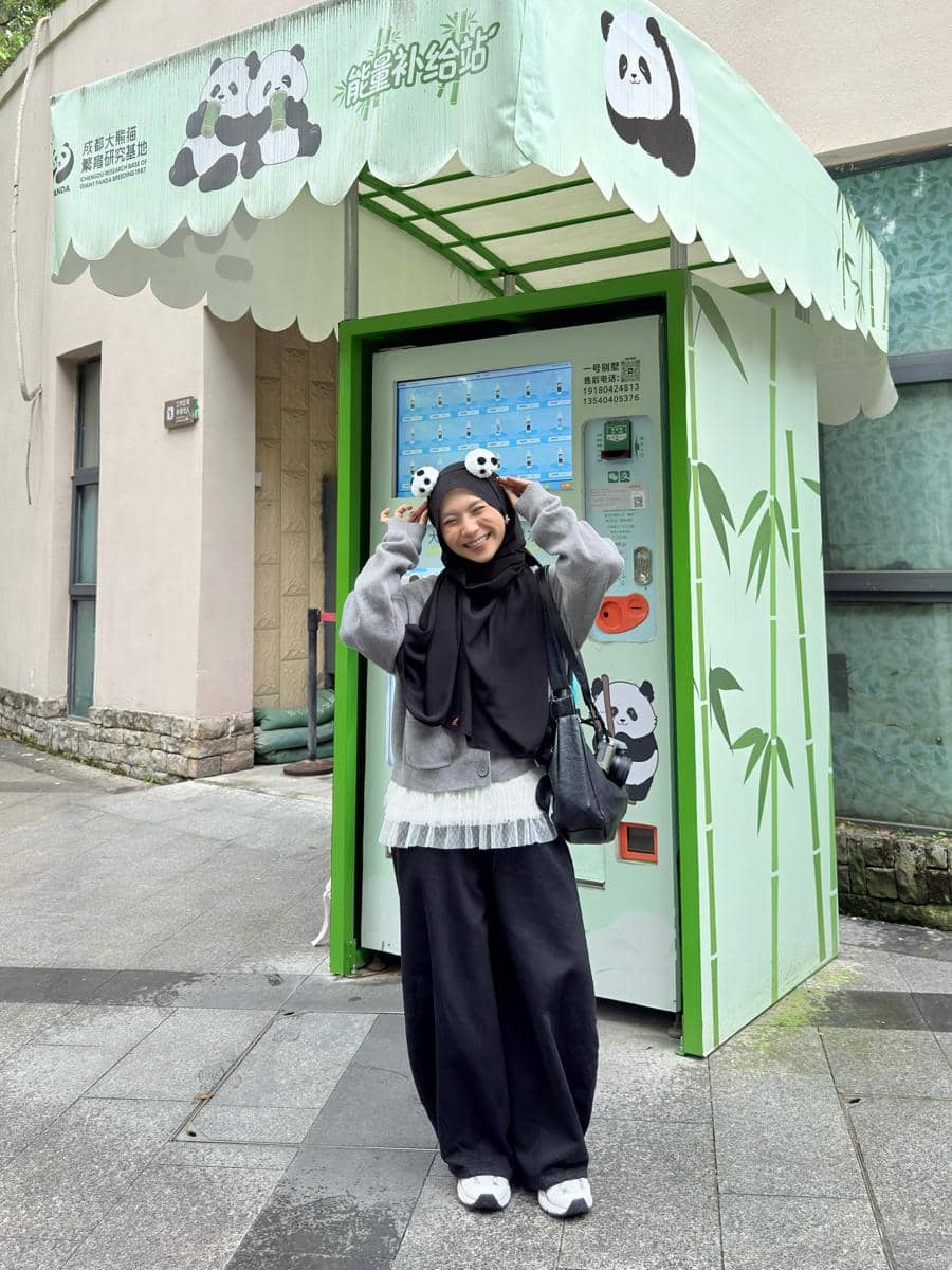 Seorang wanita berhijab bergaya kasual dengan sweater abu-abu dan celana hitam berpose ceria di depan mesin otomatis bertema panda.