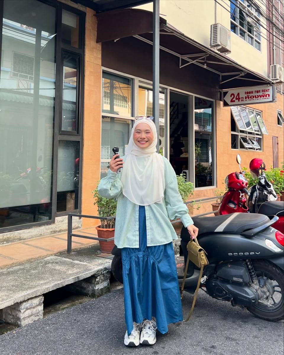 Seorang wanita berhijab mengenakan kemeja hijau muda dan rok biru panjang berpose sambil tersenyum di depan gedung dengan motor di sampingnya.