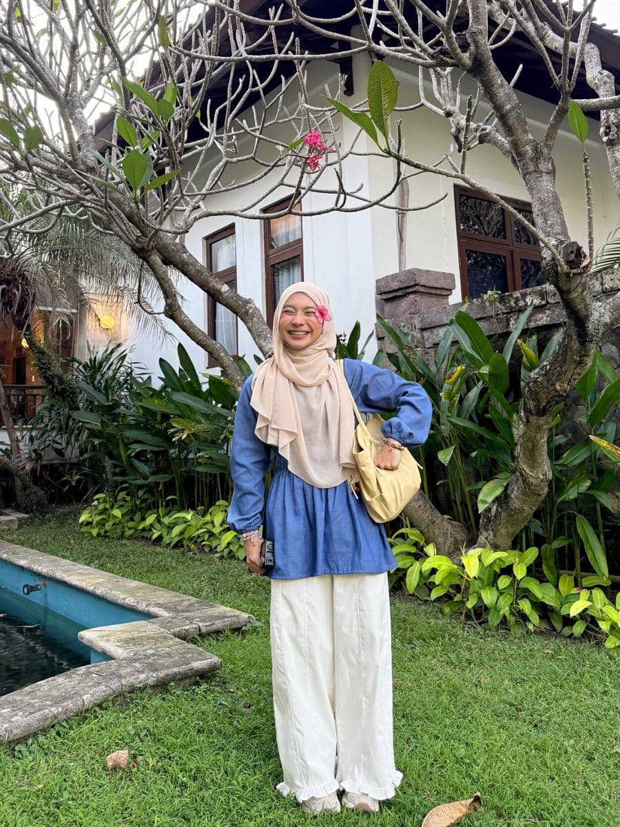 Perempuan berhijab mengenakan atasan biru dan celana putih berpose di taman hijau dengan latar rumah bergaya tropis.