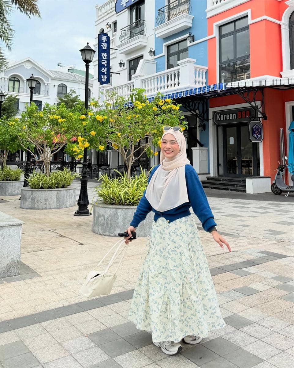 Seorang wanita berhijab tersenyum mengenakan cardigan biru dan rok panjang bermotif bunga sambil berjalan di area kota dengan bangunan berwarna cerah.