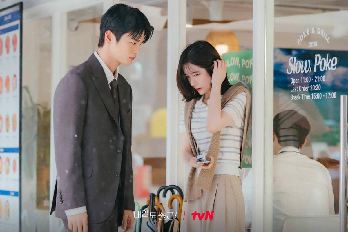 Dua karakter dalam drama See You at Work Tomorrow berdiri di depan kafe, pria mengenakan setelan jas dan wanita memegang ponsel sambil menunduk.