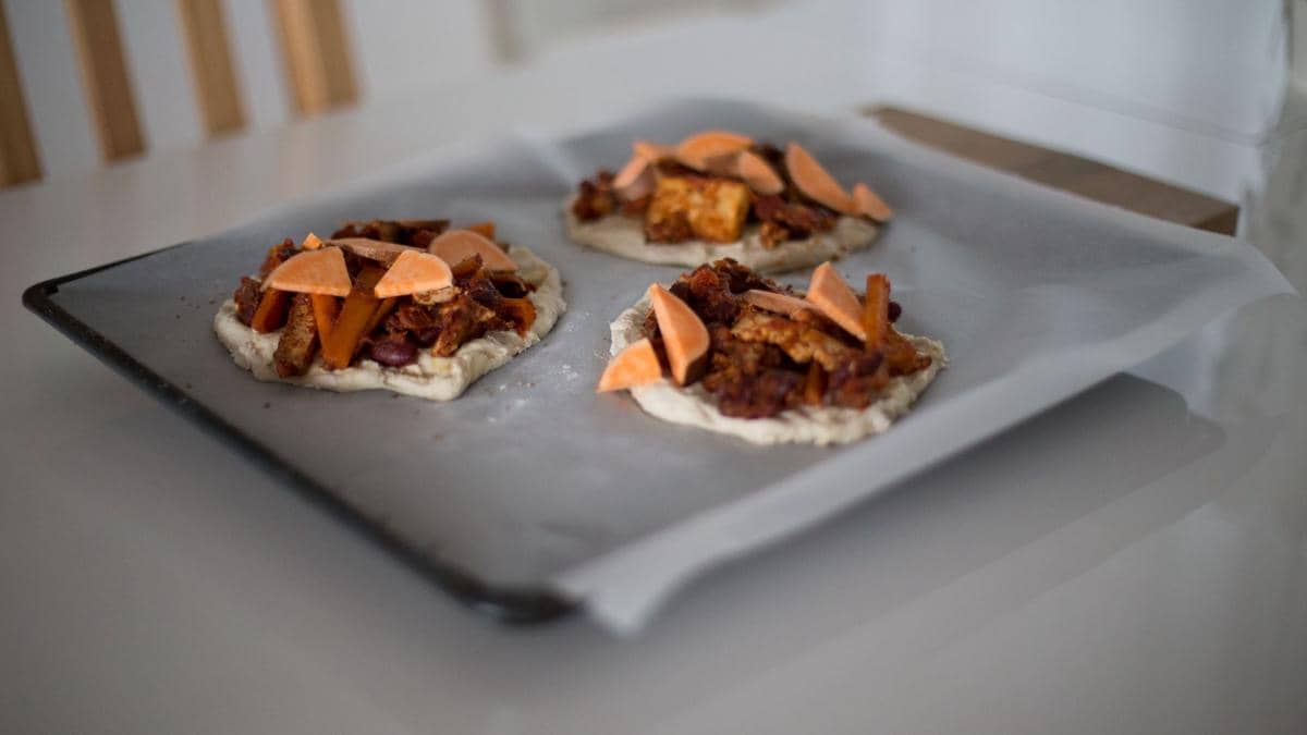 Tiga mini pizza dengan topping daging dan potongan keju oranye di atas loyang berlapis kertas roti siap dipanggang di oven.