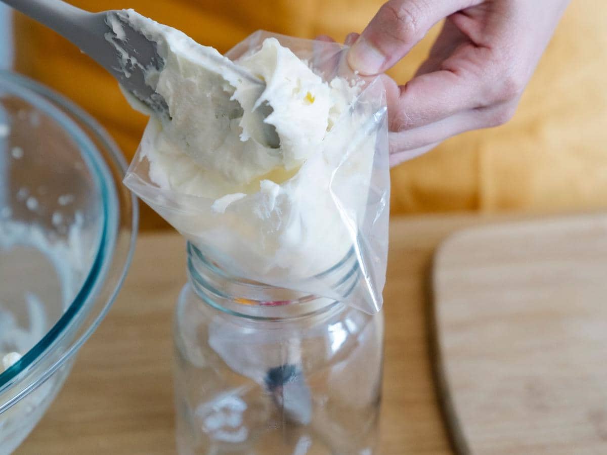 Tangan memegang spatula saat memasukkan adonan krim putih ke dalam plastik segitiga yang diletakkan di atas toples kaca.