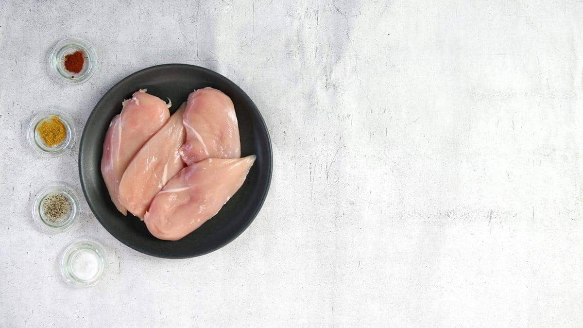 Beberapa potong dada ayam mentah di atas piring hitam dengan empat wadah kecil berisi bumbu di permukaan meja terang.