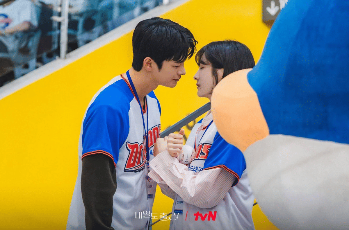 Dua karakter drama See You at Work Tomorrow mengenakan seragam bisbol biru putih saling menatap dekat di tribun stadion.