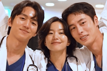 Tiga dokter muda mengenakan jas putih dan seragam medis biru tersenyum bersama di rumah sakit dalam cuplikan drama Korea Doctor on the Edge.