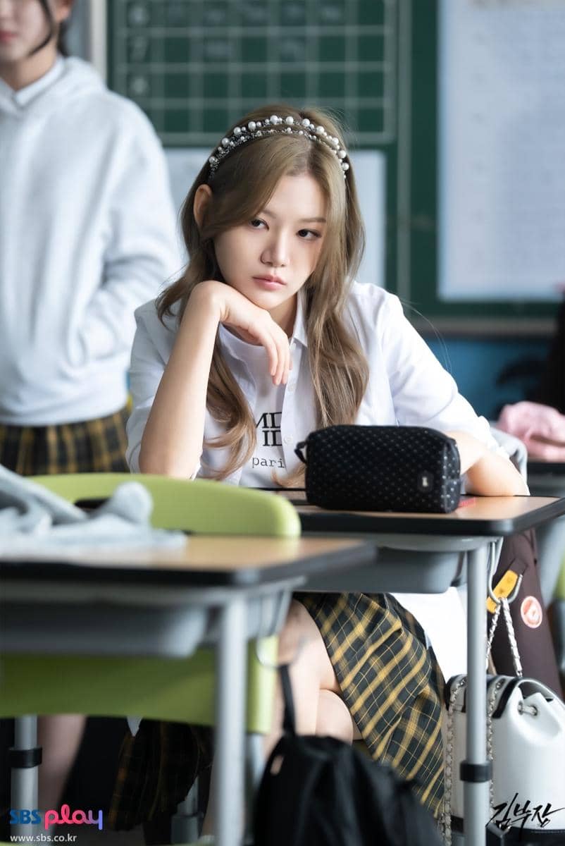 Seorang siswi berseragam duduk di bangku kelas dengan ekspresi serius, mengenakan bando mutiara dan kemeja putih di lokasi syuting drama Agent Kim Reactivated.