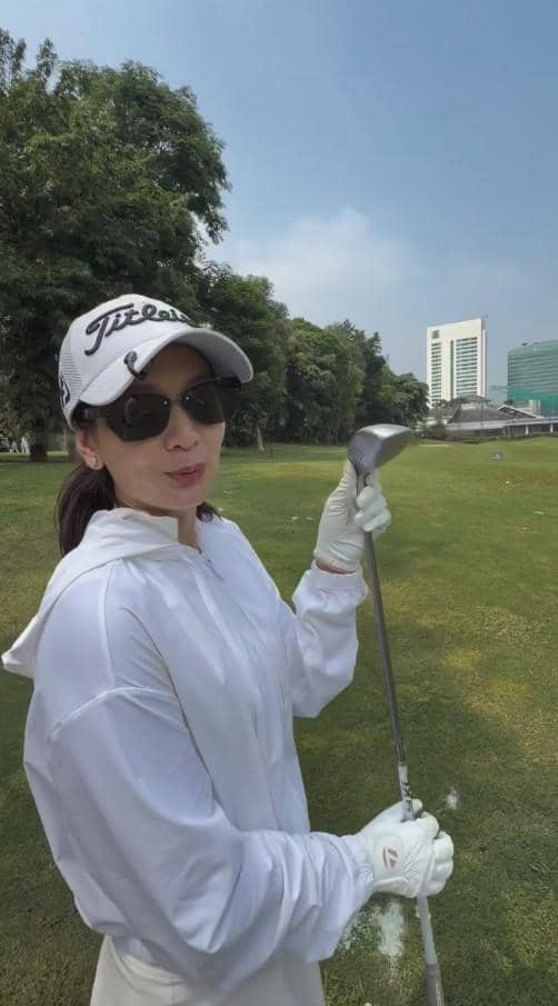 Seorang perempuan mengenakan pakaian putih, topi, dan kacamata hitam sedang memegang stik golf di lapangan hijau dengan gedung di latar belakang.