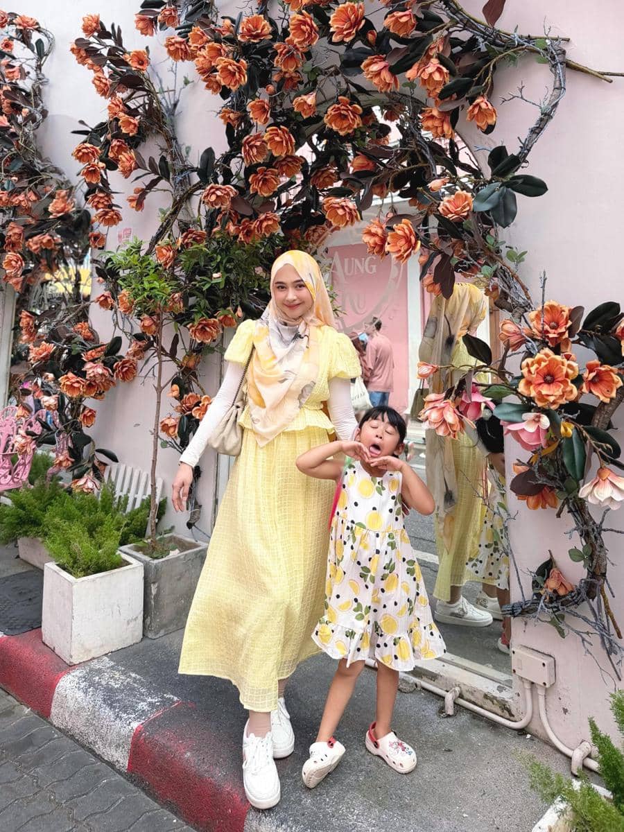 Seorang wanita berhijab dan anak kecil berpose ceria di depan dekorasi bunga oranye di area luar ruangan dengan suasana cerah.