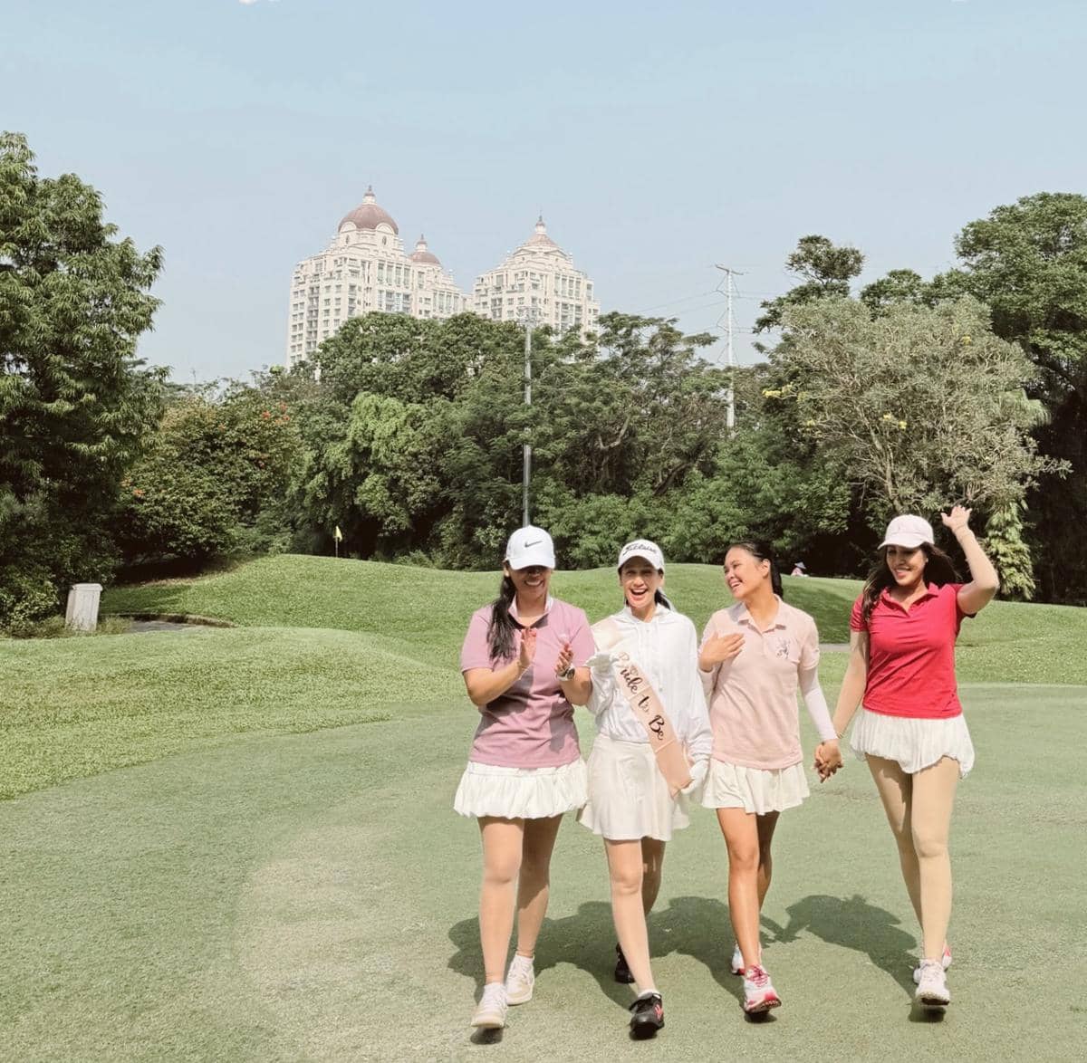 Empat wanita mengenakan pakaian golf berwarna cerah berjalan bersama di lapangan hijau dengan latar gedung tinggi dan pepohonan.