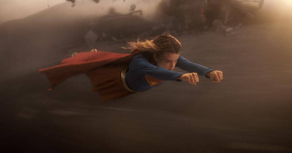 Supergirl terbang cepat di udara dengan latar belakang gelap dan puing-puing, mengenakan kostum biru dan jubah merah khasnya.