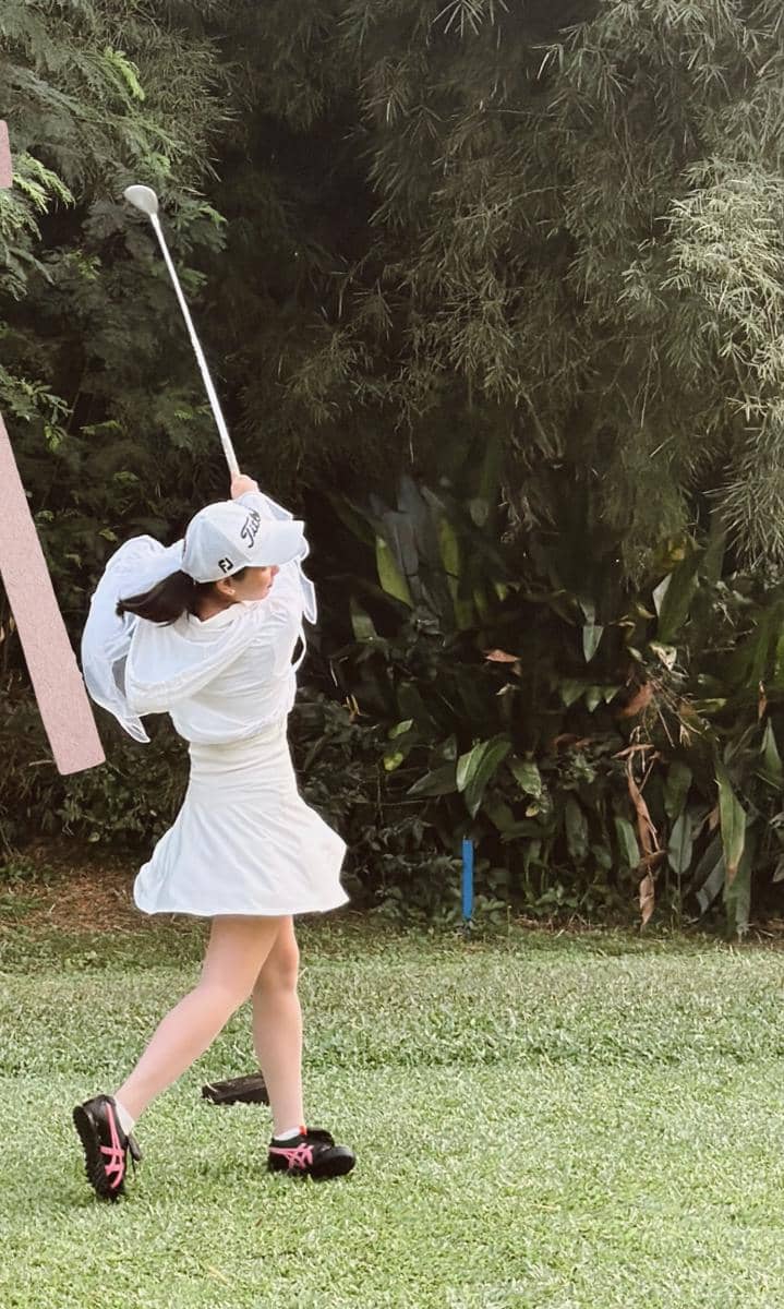 Seorang perempuan mengenakan gaun putih dan topi golf sedang mengayunkan stik golf di lapangan hijau dengan latar pepohonan.